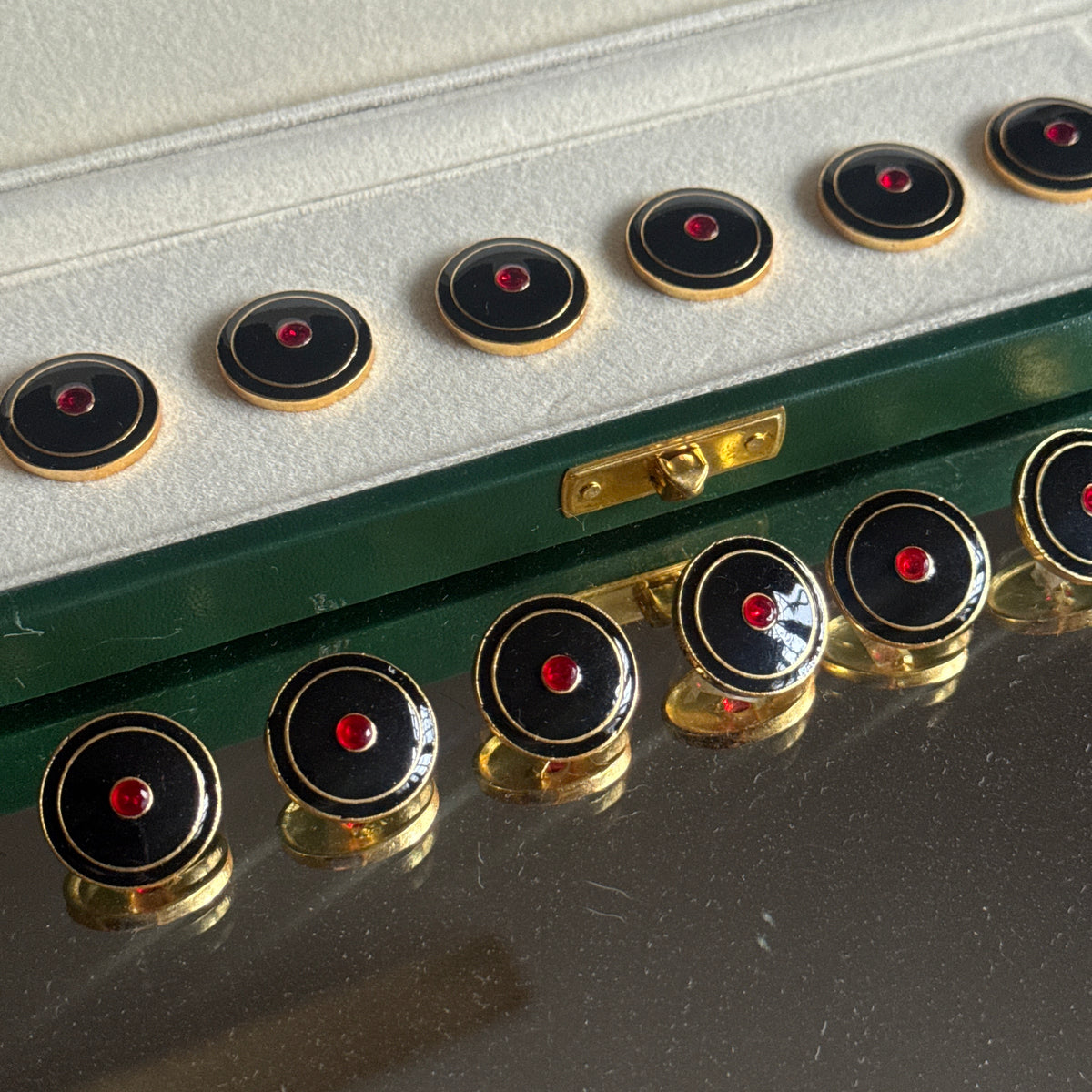 A set of round black enamel buttons with gold trim and a small red center stone, shown both inside an open green presentation box and arranged on a reflective dark surface.