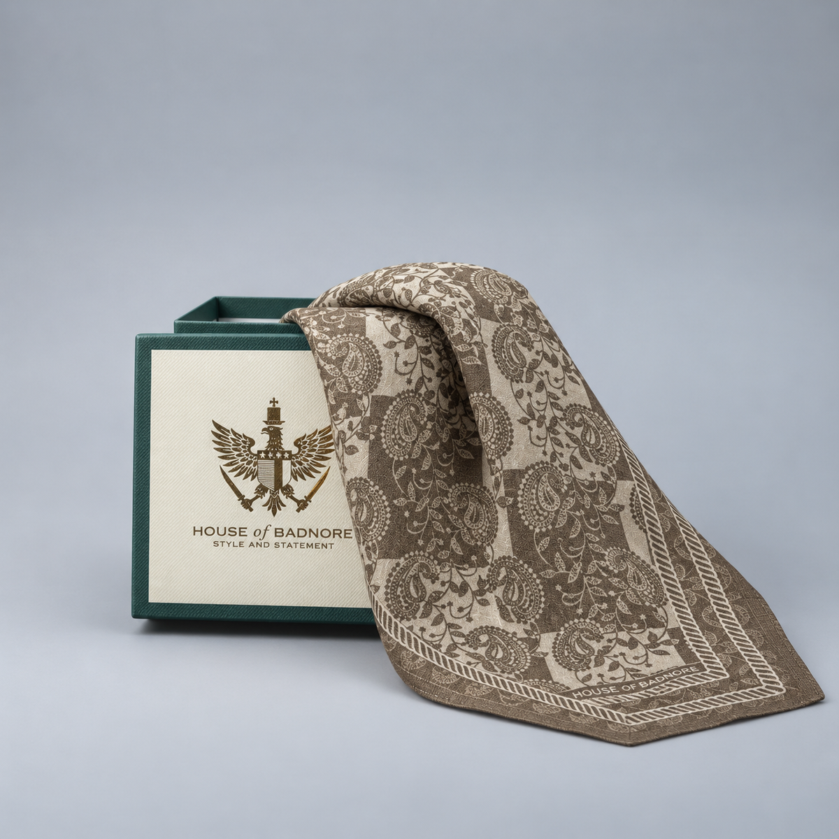 A square patterned scarf in ivory and beige with paisleys draped over a branded green and cream presentation box, photographed on a clean light gray studio background.