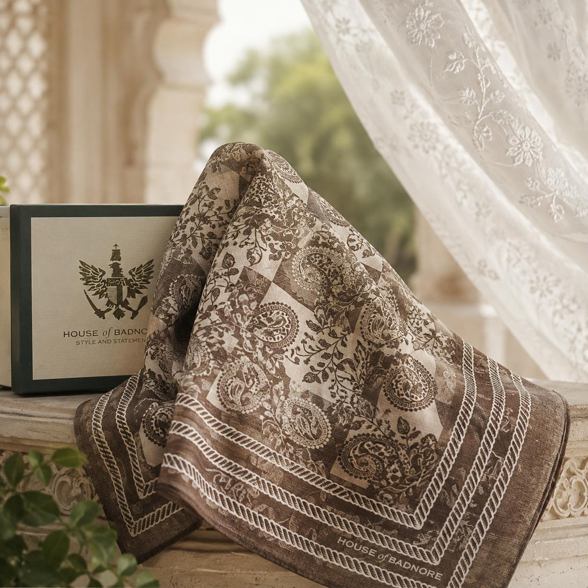 an ivory and beige patterned scarf with paisleys draped over a stone ledge next to its cream presentation box, with a white lace curtain and soft-focus outdoor background.