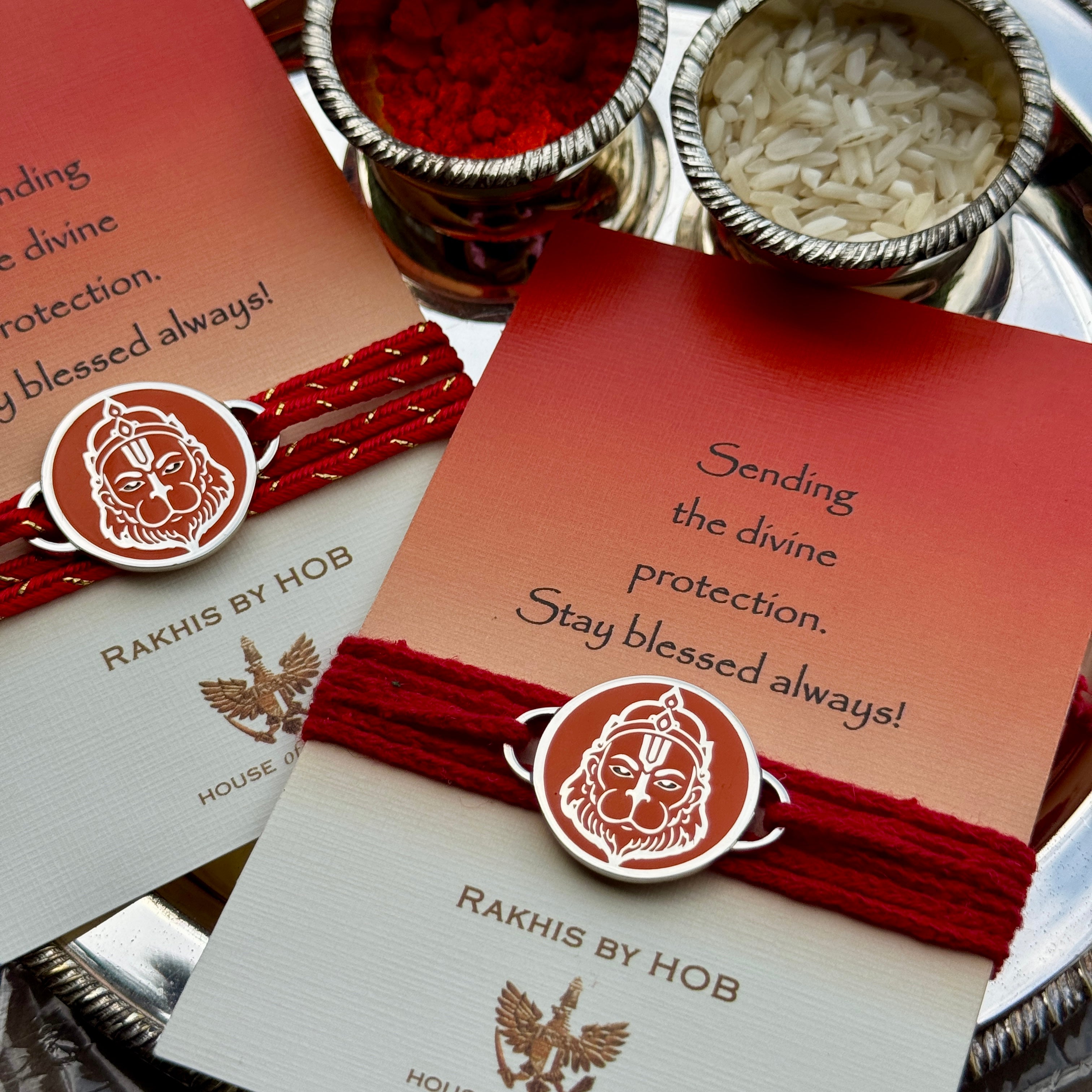 A close-up of a silver-toned Rakhi bracelet with a circular Hanuman emblem, wrapped around a red cord and attached to a red-to-cream card.