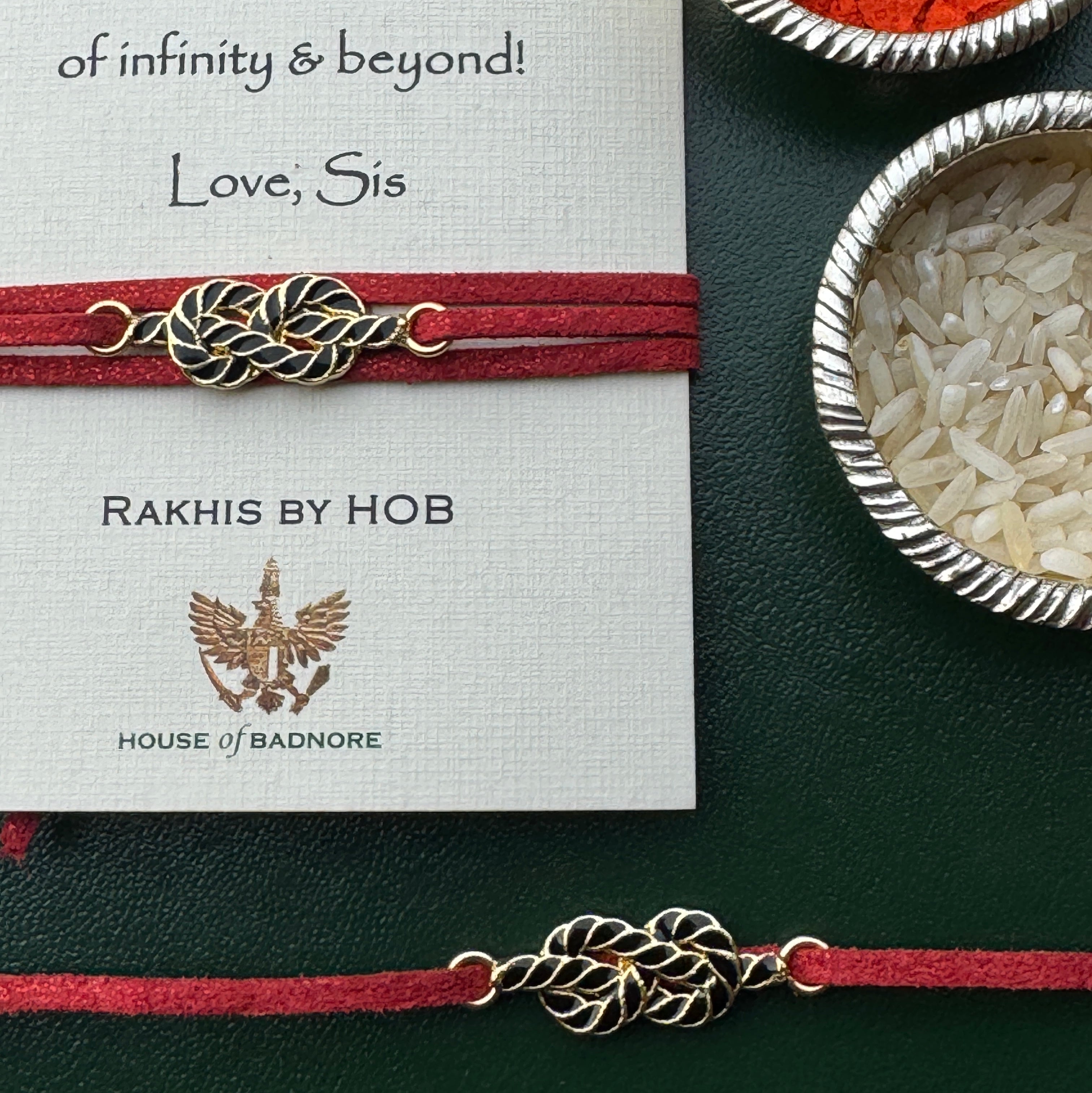 A red fabric rakhi bracelet with a detailed gold and black-toned braided knot charm, displayed on a white gift card from 'Rakhis by Hob' and 'House of Badnore'. The card is styled on a dark green textured surface alongside bowls of rice and red powder.