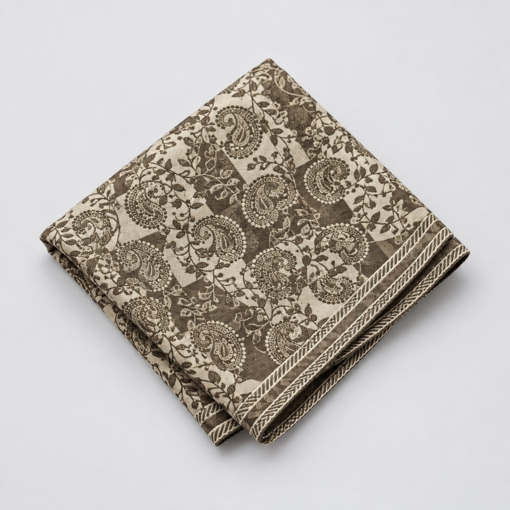 A close-up of a square textile with an intricate paisley and floral pattern in dark beige and ivory, featuring a decorative braided border.