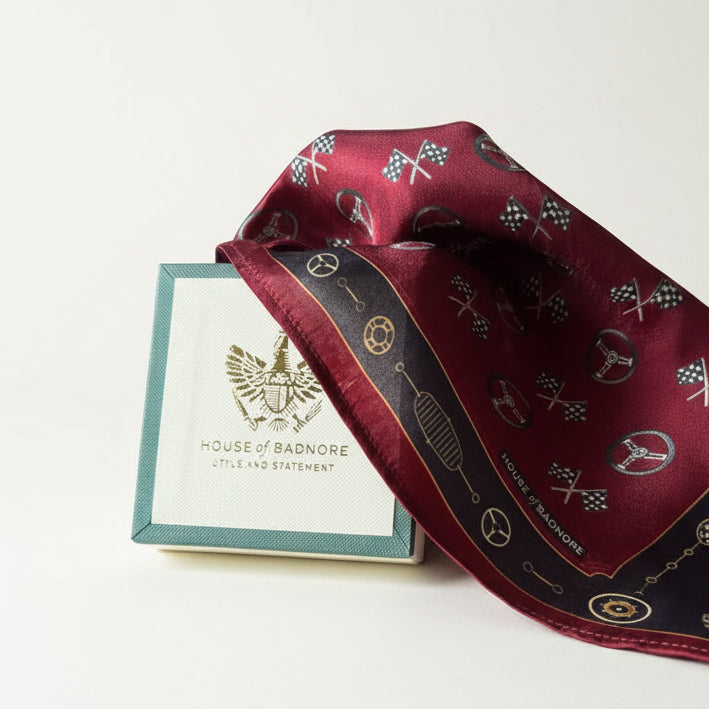 A maroon silk scarf with a black border and gold automotive-themed icons, displayed next to its light blue and white House of Badnore gift box.