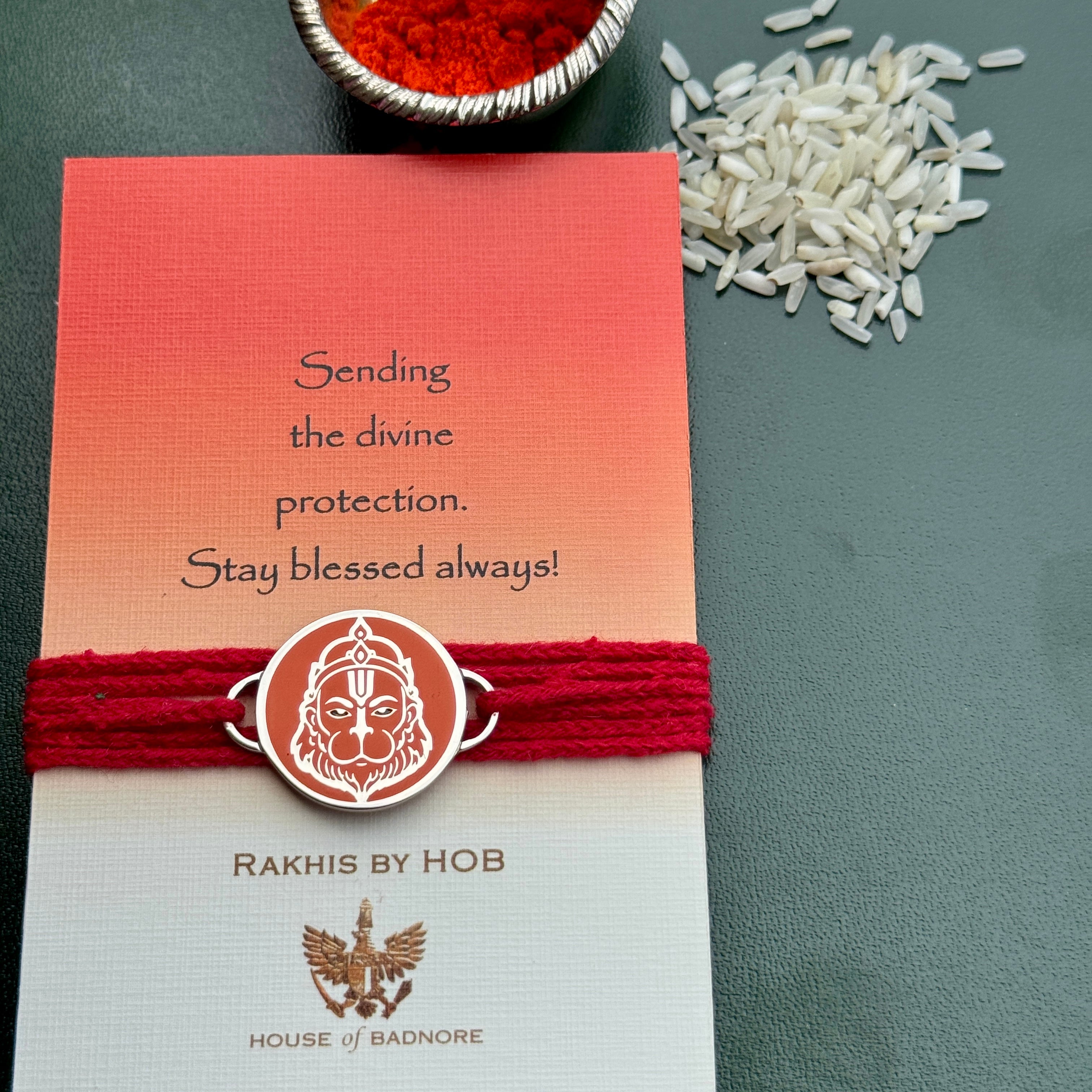 A red and orange gradient Rakhi card with a red thread bracelet and a circular Hanuman emblem, styled on a dark green surface with rice grains and a bowl of red powder.