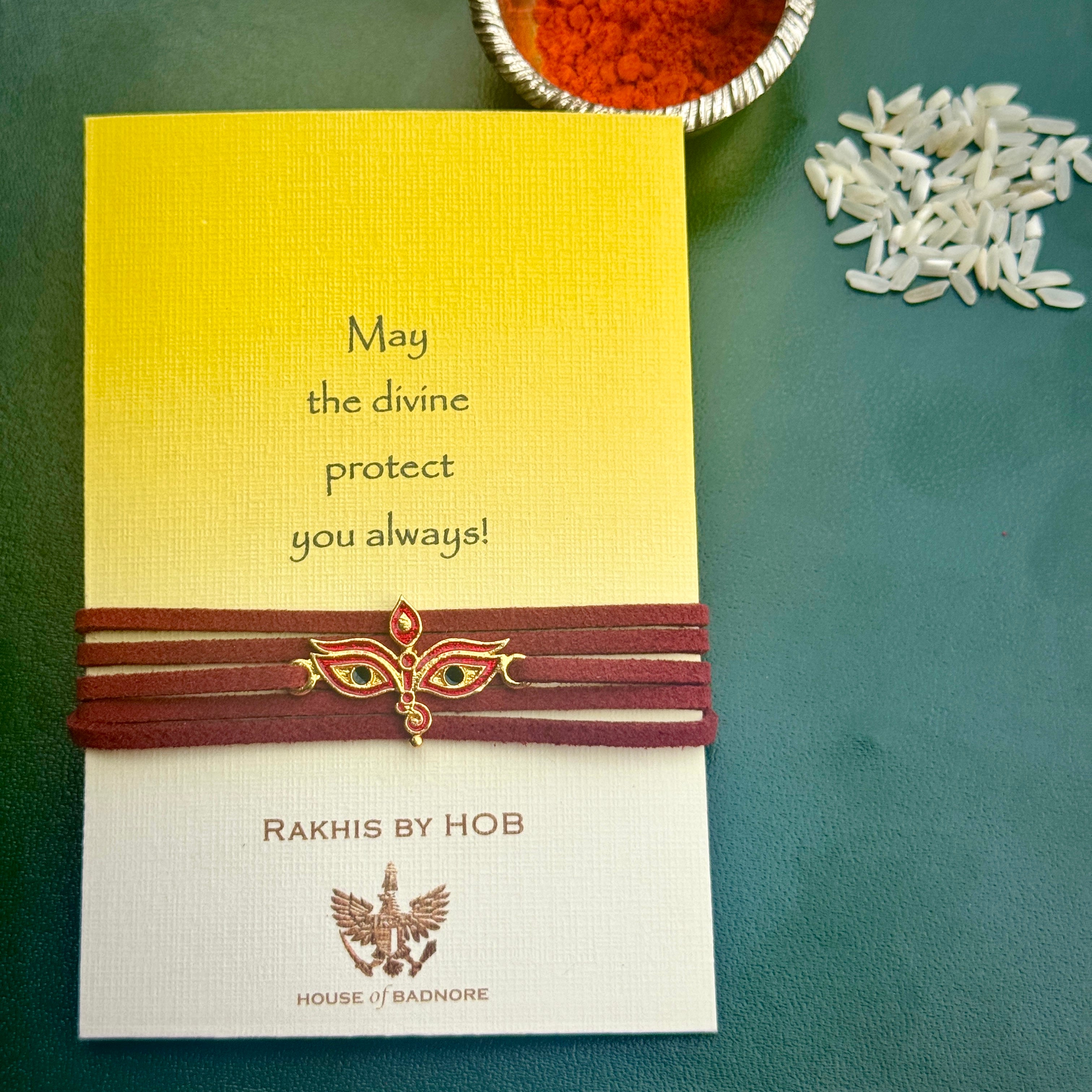 A Rakhi presented on a branded card. The Rakhi has a maroon cord with the central in the shape of a woman's eyes.