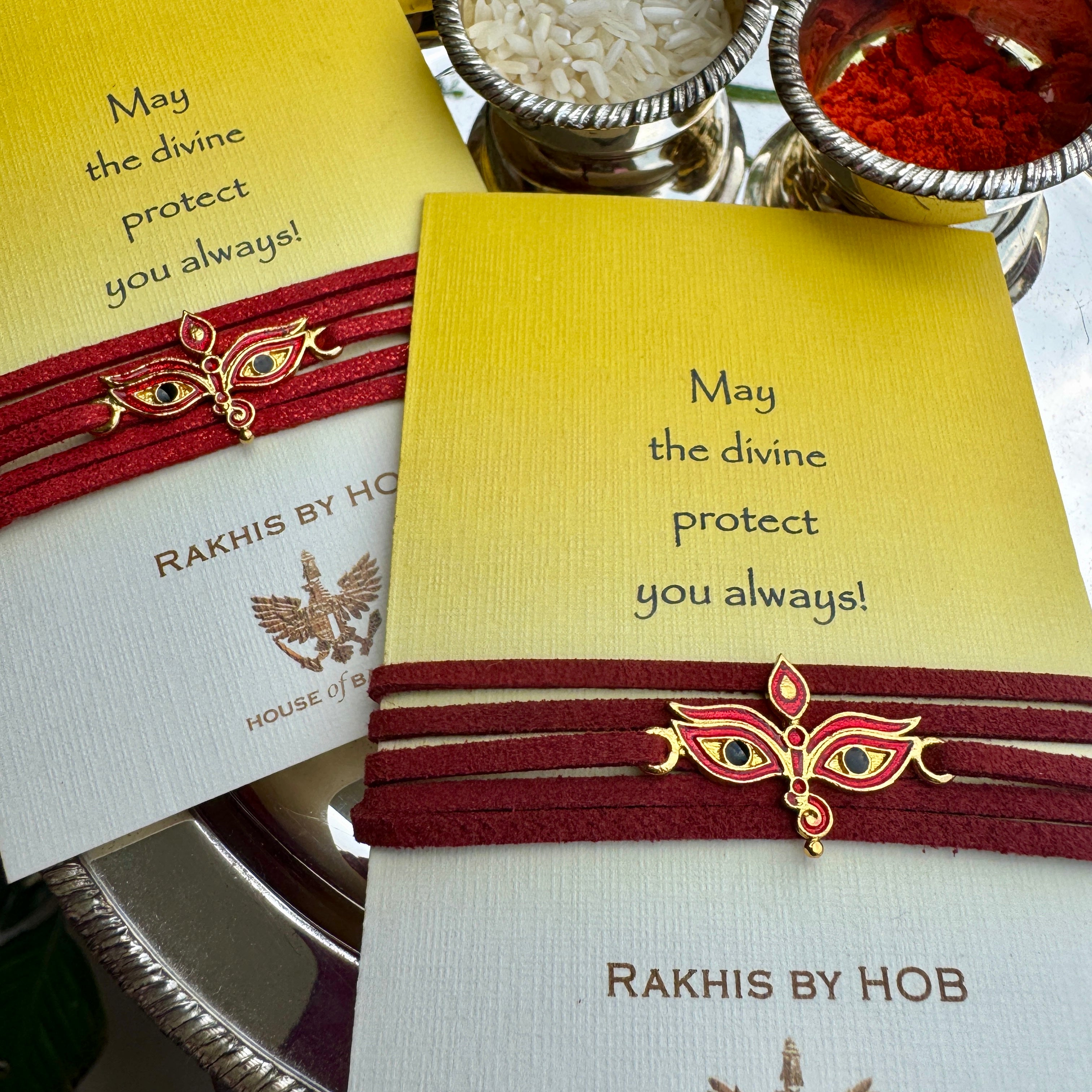 A close-up of a Rakhi bracelet presented on its branded card. The Rakhi central motif are a woman's eyes, wrapped in dark red cord, and displayed on a white and yellow card.