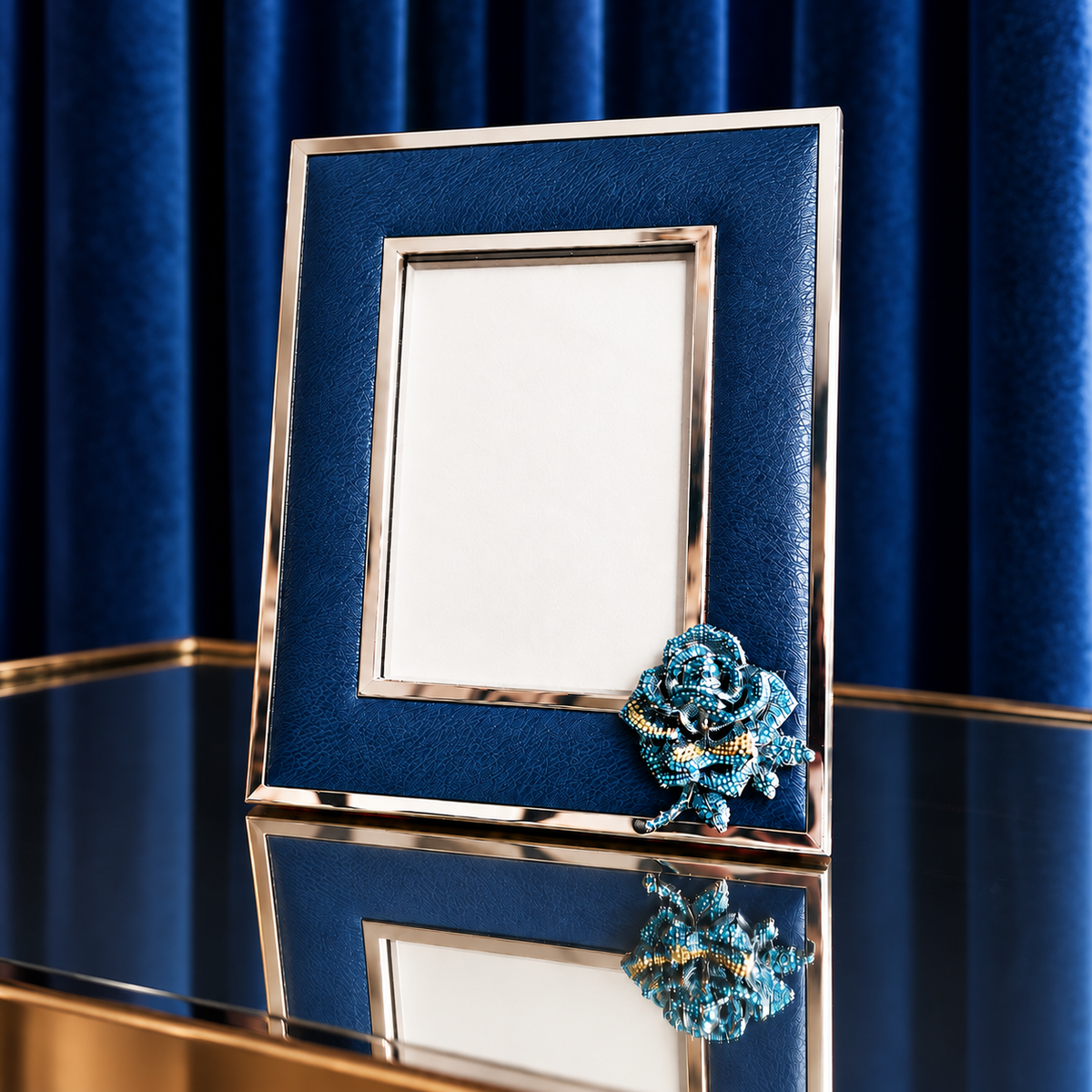 A navy blue and silver picture frame with a blue rose embellishment in the bottom right corner, set on a reflective surface against a dark blue velvet curtain background.