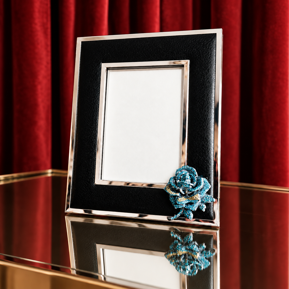 A black and silver picture frame with a decorative blue rose embellishment on the lower right corner, displayed on a reflective gold table against a red velvet curtain background.