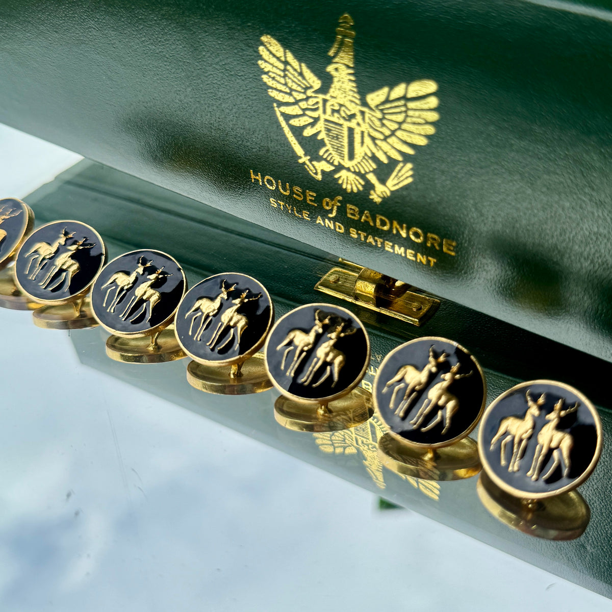 A close-up of round black buttons with gold stag motifs, displayed on a matching dark green presentation box embossed with the House of Badnore logo.